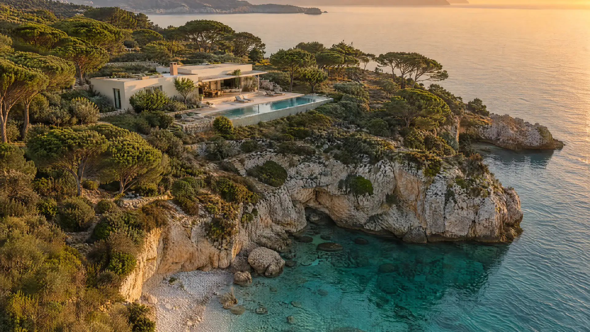 Vue aérienne d’une villa de pierre parmi les pins, au-dessus d’une crique turquoise, sur la presqu’île fictive de Maison Sérane.